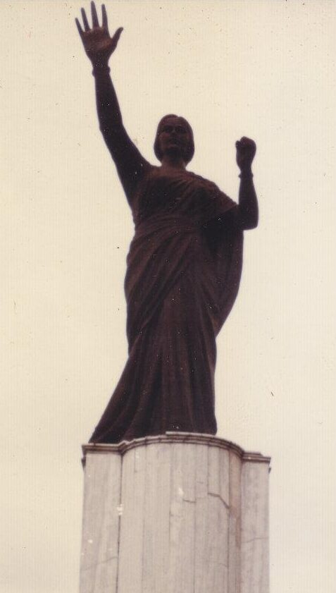 A dark full-length statue of a woman in a sari with one arm raised, mounted on a tall fluted pedestal against a pale sky.