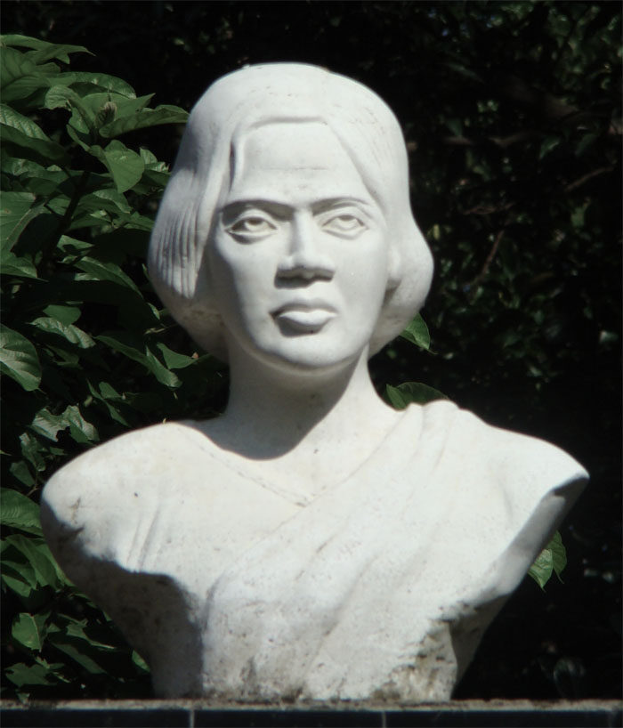 A white stone bust of Pritilata Waddedar with jaw-length hair and a draped sari, against dark green foliage.