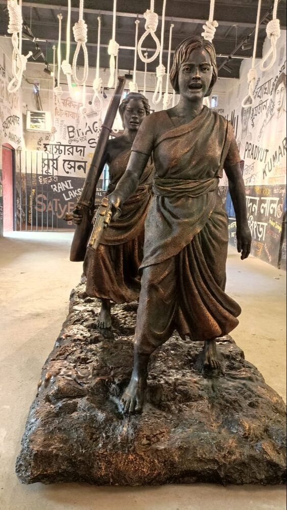 A bronze-toned sculpture of two women in saris striding forward in a museum gallery, the walls behind them painted with the names of revolutionaries.