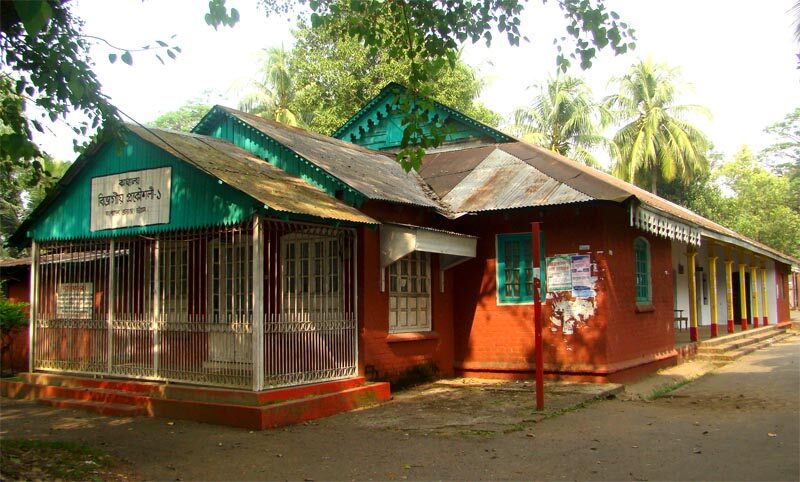 A single-storey colonial-period building with a green corrugated roof and red walls, among trees.