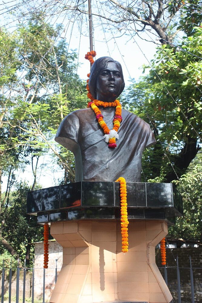 A dark bronze bust of Pritilata Waddedar on a stepped plinth, hung with marigold garlands, outdoors among trees.